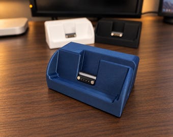 May include: A blue charging dock featuring a recessed slot and a four-pin magnetic connector. The dock has a slanted front face designed to hold a device at an angle. Two additional charging docks in white and black are visible in the background.