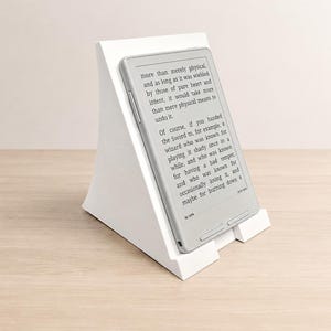 May include: A white, angled e-reader stand holding a light grey e-reader. The screen displays text starting with "more than merely physical," and includes a battery icon with "at 100%" text at the bottom. The stand has a solid, triangular profile.