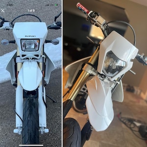 Peut inclure: Une moto Suzuki blanche avec un phare noir et un garde-boue blanc. La moto a une fourche avant dorée et un guidon noir.