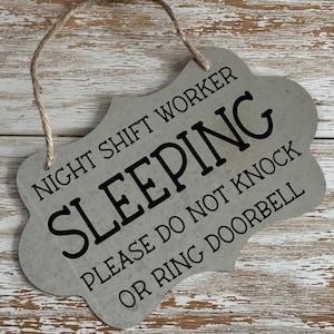 May include: A gray metal sign with rounded edges and a string for hanging. The sign reads "Night Shift Worker Sleeping Please Do Not Knock Or Ring Doorbell".