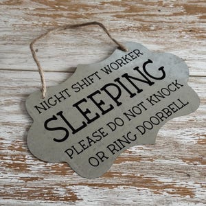 May include: A metal sign with a distressed silver finish and black text that reads "Night Shift Worker Sleeping Please Do Not Knock Or Ring Doorbell". The sign has a rope loop for hanging.