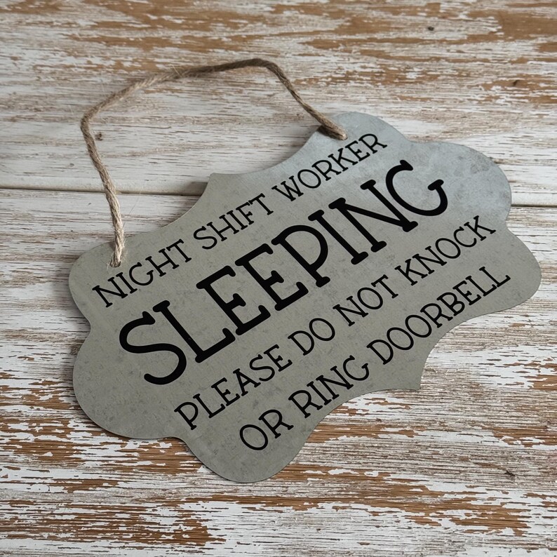 May include: A metal sign with a distressed silver finish and black text that reads "Night Shift Worker Sleeping Please Do Not Knock Or Ring Doorbell". The sign has a rope loop for hanging.