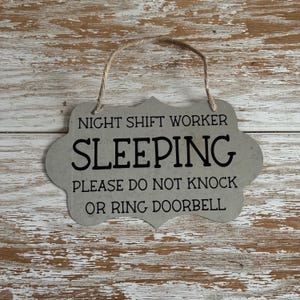 May include: A gray sign with a rope hanger reads "Night Shift Worker Sleeping Please Do Not Knock Or Ring Doorbell".