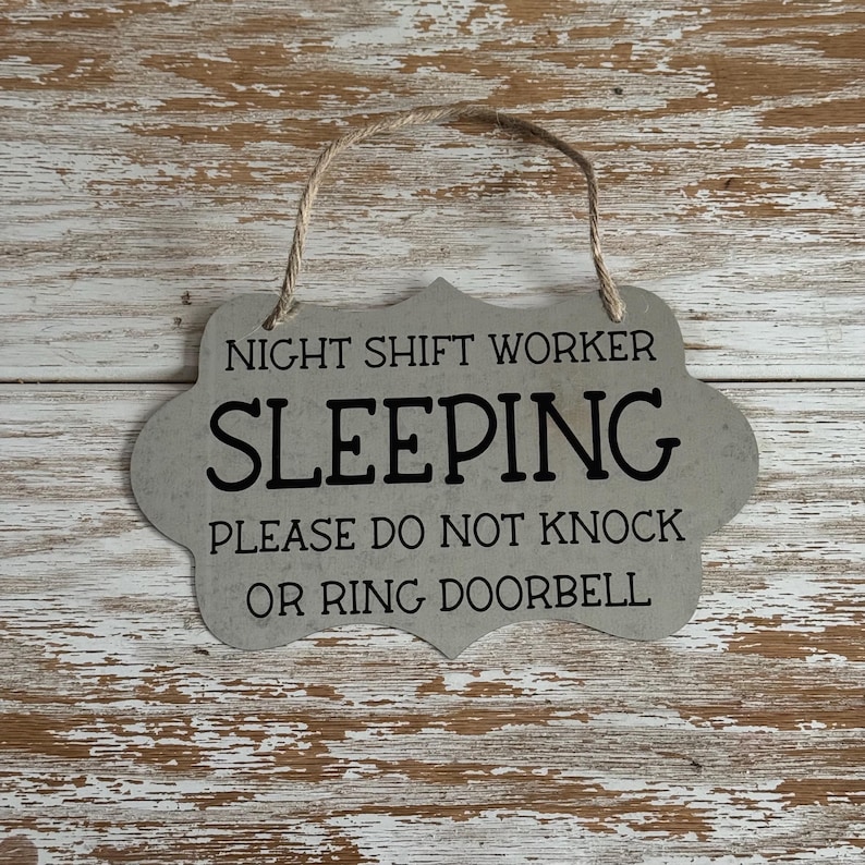May include: A gray sign with a rope hanger reads "Night Shift Worker Sleeping Please Do Not Knock Or Ring Doorbell".