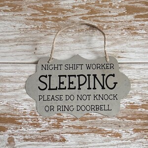 May include: A gray metal sign with a rope hanger. The sign reads "Night Shift Worker Sleeping Please Do Not Knock Or Ring Doorbell".