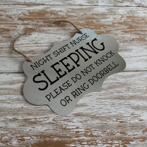 May include: A silver metal sign with a decorative edge and a jute string for hanging. The sign reads "NIGHT SHIFT NURSE SLEEPING PLEASE DO NOT KNOCK OR RING DOORBELL" in black text.