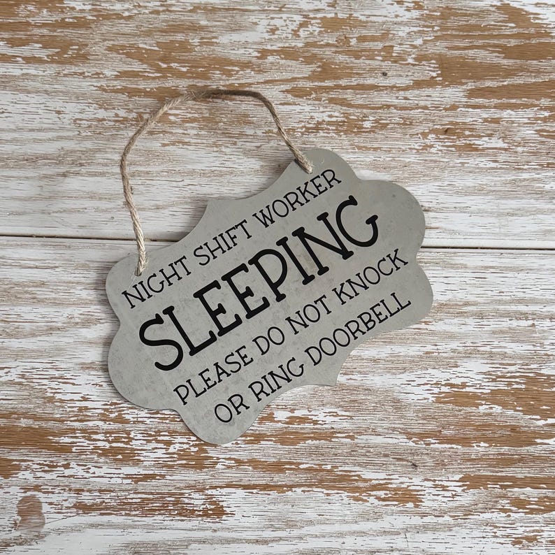 May include: A silver metal sign with a rope hanger reads "Night Shift Worker Sleeping Please Do Not Knock Or Ring Doorbell".