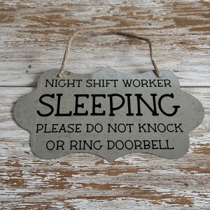 May include: A gray metal sign with a rustic finish and a rope hanger. The sign reads "Night Shift Worker Sleeping Please Do Not Knock Or Ring Doorbell".