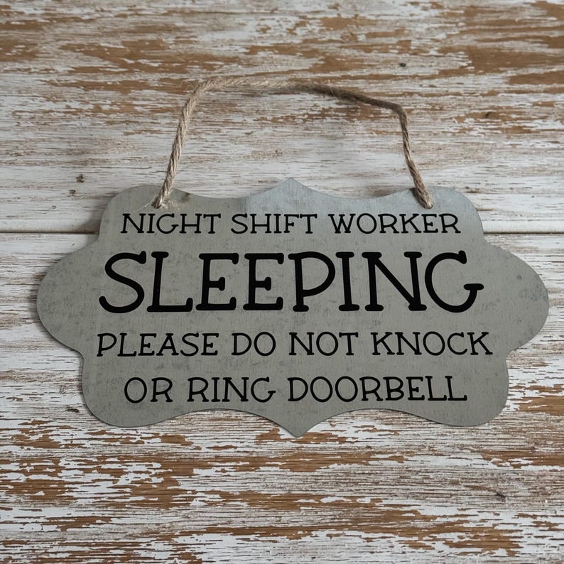 May include: A gray metal sign with a rustic finish and a rope hanger. The sign reads "Night Shift Worker Sleeping Please Do Not Knock Or Ring Doorbell".
