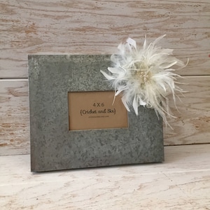 May include: A galvanized metal picture frame with a white feather flower embellishment. The frame is designed to hold a 4x6 inch photo and has the text "4x6 (Cricket and Six) cricketandsix.etsy.com" printed on the mat.