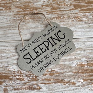 May include: A silver metal sign with a distressed finish and black text that reads "Night Shift Worker Sleeping Please Do Not Knock Or Ring Doorbell". The sign has a hole at the top for hanging.