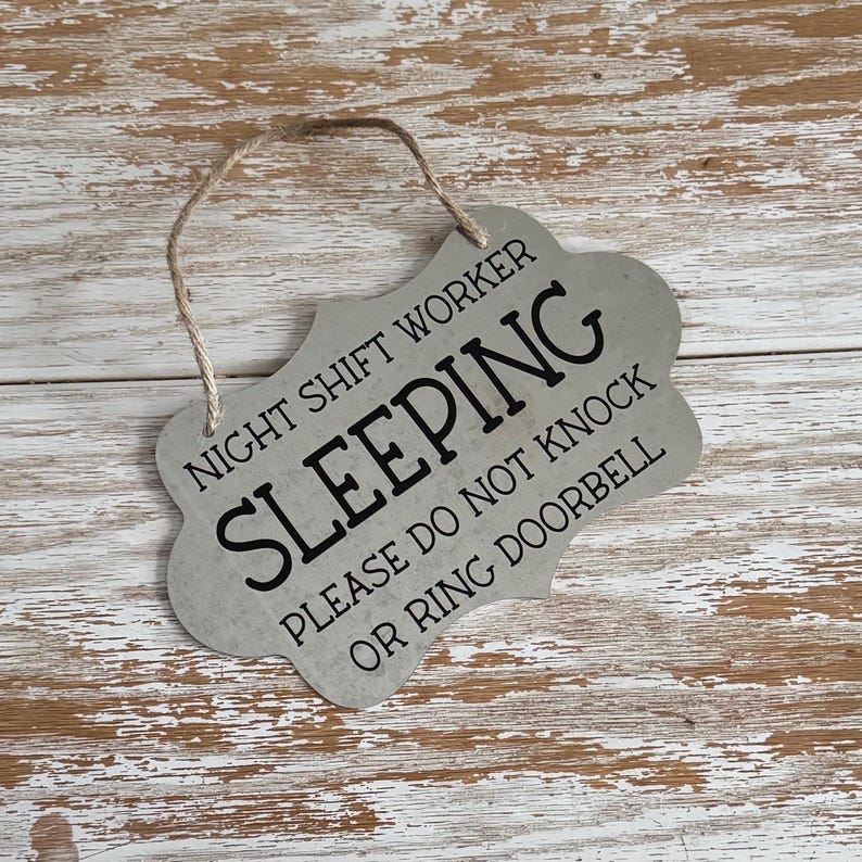 May include: A silver metal sign with a distressed finish and black text that reads "Night Shift Worker Sleeping Please Do Not Knock Or Ring Doorbell". The sign has a hole at the top for hanging.