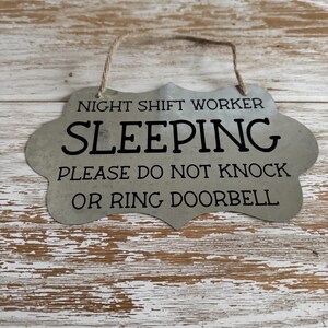 May include: A silver metal sign with a rustic finish and a rope hanger. The sign reads "Night Shift Worker Sleeping Please Do Not Knock Or Ring Doorbell".