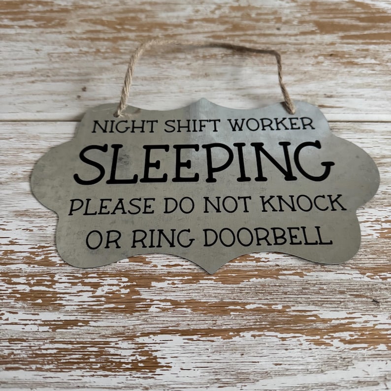 May include: A silver metal sign with a rustic finish and a rope hanger. The sign reads "Night Shift Worker Sleeping Please Do Not Knock Or Ring Doorbell".