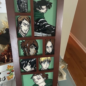 Op de afbeelding: Een ingelijst kunstwerk met een collage van anime-personages uit de populaire serie Attack on Titan. De personages zijn afgebeeld in een glas-in-loodstijl met levendige kleuren en ingewikkelde details. De lijst is van bruin hout met een groene achtergrond.