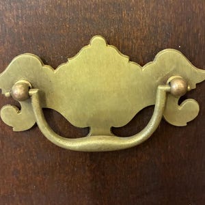 Könnte beinhalten: Ein Messing-Schubladengriff mit einem verzierten Design. Der Griff hat einen gebogenen Griff und eine dekorative Rückplatte mit einer symmetrischen, stilisierten Form. Die Hardware ist auf einer dunklen Holzoberfläche angebracht.