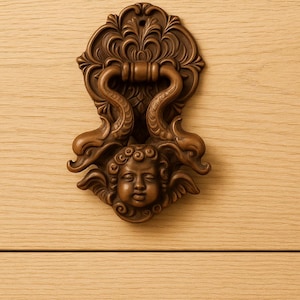 May include: A detailed, brown, antique-style door knocker. The knocker features an ornate design with a cherub face and scrolled accents. The handle is a curved bar, and the knocker is mounted on a light wood surface.