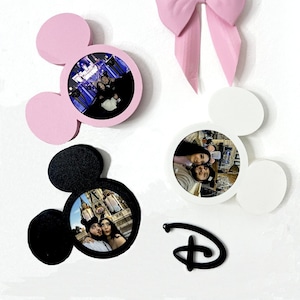 Op de afbeelding: Drie Mickey Mouse-vormige fotolijsten in roze, zwart en wit, met een zwart Disney-logo. Elk frame heeft een ronde opening voor een foto. De frames hangen aan een witte muur.