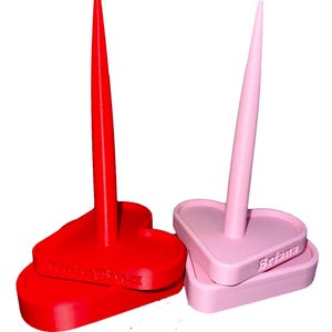 May include: Two heart-shaped stands with a pointed spike in the center. The stand on the left is red and the stand on the right is pink. Both stands have the word "Stylus" printed on the bottom.