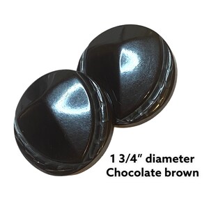 Puede incluir: Dos botones marrón chocolate, cada uno de 4,4 cm de diámetro, sobre un fondo blanco. Los botones tienen un acabado brillante y un diseño texturizado en los bordes. El texto "1 3/4" diameter Chocolate brown" es visible.
