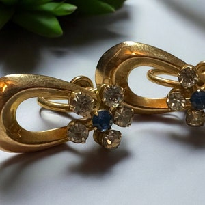 Könnte beinhalten: Ein Paar goldfarbene Vintage-Ohrringe. Jeder Ohrring hat ein tropfenförmiges Design mit einem floralen Cluster aus klaren und blauen Strasssteinen. Die Ohrringe haben einen klassischen, eleganten Stil.