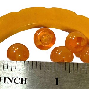 May include: Orange-colored, translucent, domed beads with a flat back, next to a ruler showing inches. The beads are approximately 0.5 inches in diameter. A curved, orange-colored object is in the background. The beads have a glossy finish.