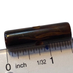 May include: A cylindrical, brown object with a marbled appearance, approximately 1 inch long. The object is smooth and appears to be made of a solid material. A ruler is placed next to the object for scale.