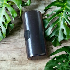 Puede incluir: Un objeto rectangular de madera marrón oscuro con un área empotrada en un lado. El objeto se encuentra sobre un fondo de hojas tropicales verdes y una superficie de madera envejecida.
