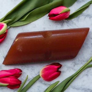 Puede incluir: Una pastilla de jabón marrón, geométrica, con una pequeña hendidura, sobre una superficie de mármol. Tulipanes rojos brillantes con tallos y hojas verdes están dispuestos alrededor del jabón.