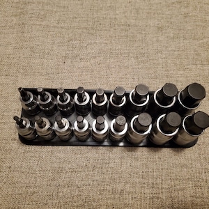 May include: A set of black and silver socket wrenches arranged in a black holder. The tools are of varying sizes and shapes, designed for mechanical work. The background is a textured, tan surface.