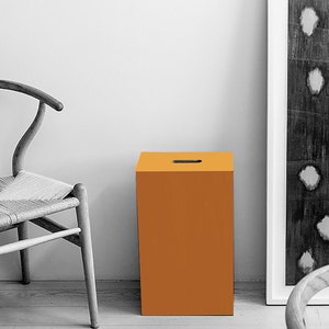 May include: A small, orange, square stool with a cutout on the top. The stool is in a room with a white wall and a wooden floor.