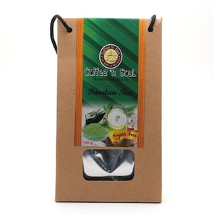 Può includere: Un sacchetto di carta marrone con manico in corda nera contenente tè al pandan. L'etichetta riporta il testo Coffee 'n Soul, Pandan Tea e Sugar Free, con un'immagine di tè verde in polvere e un bicchiere di tè. Il peso è indicato come 100 gr.