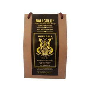 Puede incluir: Una bolsa de papel marrón con una etiqueta dorada que dice "Bali Gold* Gourmet Coffee" y "Kopi Biji/Roasted Bean". La etiqueta también presenta un logotipo de mariposa y el texto "Kopi Bali" con un símbolo de marca registrada. La bolsa está atada con una cuerda marrón.