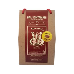 Puede incluir: Una bolsa de papel marrón de café gourmet Bali Kintamani. La bolsa presenta texto en indonesio e inglés, incluyendo "Kopi Bali" y "Single Origin 1935". También es visible un logotipo de mariposa. La bolsa tiene un asa de cordón rojo.