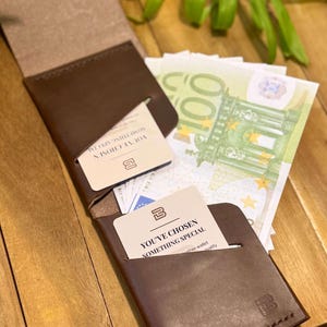 May include: A brown leather wallet with a snap closure, open to reveal a card that reads "YOU'VE CHOSEN SOMETHING SPECIAL." The wallet is next to a stack of Euro banknotes. The wallet has a stitched edge and a logo.