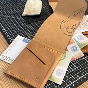 Peut inclure: Portefeuille en cuir marron clair ouvert, révélant des billets de banque en euros. Le portefeuille présente une bordure cousue et un logo en relief. L'inscription "MADE IN ITALY 1982" est estampillée à l'intérieur.