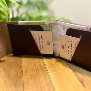 May include: Open brown leather wallet with two card slots and a snap closure. The wallet contains cards with the text "YOU'VE CHOSEN SOMETHING SPECIAL". The wallet is open, revealing cash inside. The wallet is on a wooden surface.