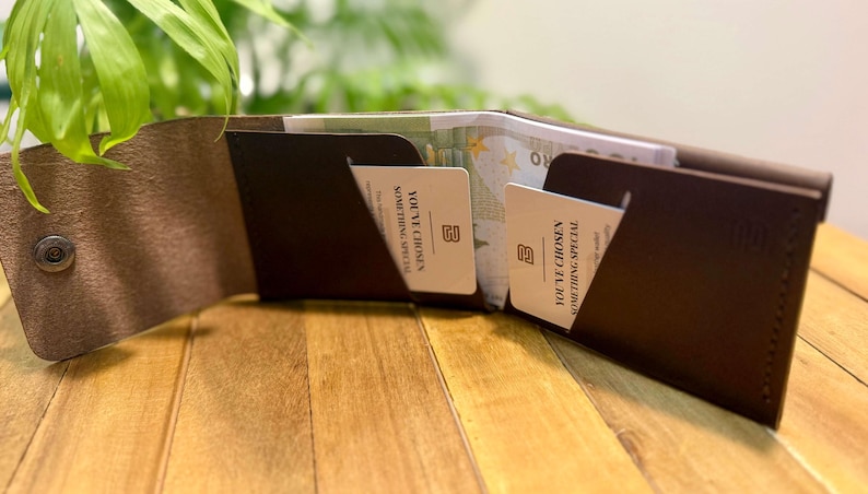 May include: Open brown leather wallet with two card slots and a snap closure. The wallet contains cards with the text "YOU'VE CHOSEN SOMETHING SPECIAL". The wallet is open, revealing cash inside. The wallet is on a wooden surface.