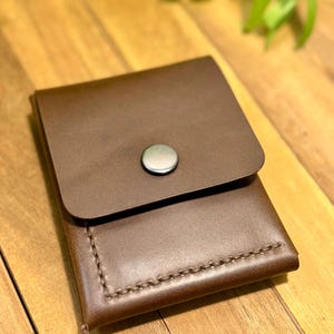 May include: A brown leather wallet with a flap closure and a silver button. The wallet is rectangular with visible stitching along the edges. The background is a wooden surface with some green foliage.