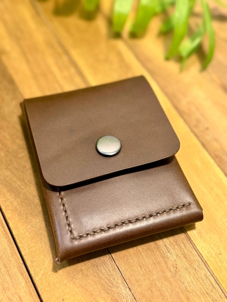 May include: A brown leather wallet with a flap closure and a silver button. The wallet is rectangular with visible stitching along the edges. The background is a wooden surface with some green foliage.