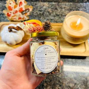 May include: A clear glass jar filled with dried botanicals and spices, topped with a gold lid, a pinecone, and dried flowers. The label reads: STOVETOP POTPOURRI SIMMER, Add all ingredients to a small pot, Add a few cups of water and simmer on low, Do not leave unattended and add water as needed, made with love.