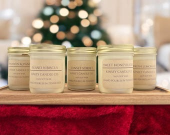 May include: Six glass candle jars with gold lids on a wooden tray. The candles are all white and have brown labels with the text "KINSEY CANDLE CO" and the scent names: "Cinnamon & Vanilla", "Island Hibiscus", "Sunset Sorbet", "Sweet Honeysuckle", and "Fresh Lemonade".