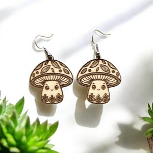 May include: A pair of wooden mushroom earrings with a cute cartoon face and leaf details. The earrings are brown and have silver hooks.