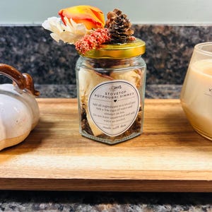 May include: A glass jar filled with dried botanicals, topped with a gold lid, a pinecone, and artificial flowers. The label reads: STOVETOP POTPOURRI SIMMER, Add all ingredients to a small pot, Add a few cups of water and simmer on low.