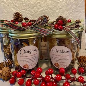 May include: Several glass jars filled with potpourri, labeled "Christmas Stovetop Potpourri Simmer." Each jar is adorned with a festive ribbon, pinecones, and red berries. The jars are arranged with a sprig of red berries in the foreground.
