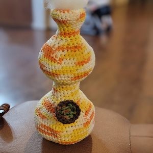 May include: A crocheted vase with a yellow, orange, and white striped pattern. The vase has a round base and a smaller, round top. A small green and brown crocheted flower is in the center of the vase.