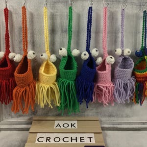 May include: A set of eight crocheted hanging toy baskets with colorful fringe. Each basket has a white crocheted eye and a crocheted mouth. The baskets are hanging from a white wooden board with the text "AOK CROCHET" on two wooden signs.