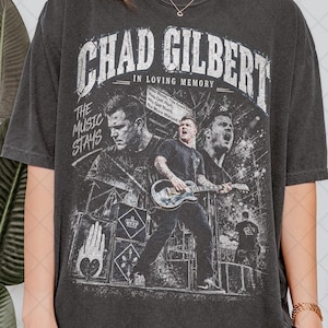 May include: A charcoal gray short-sleeve t-shirt featuring a distressed white graphic print of Chad Gilbert. The design includes the text "CHAD GILBERT IN LOVING MEMORY" and "THE MUSIC STAYS" with images of a musician playing an electric guitar.