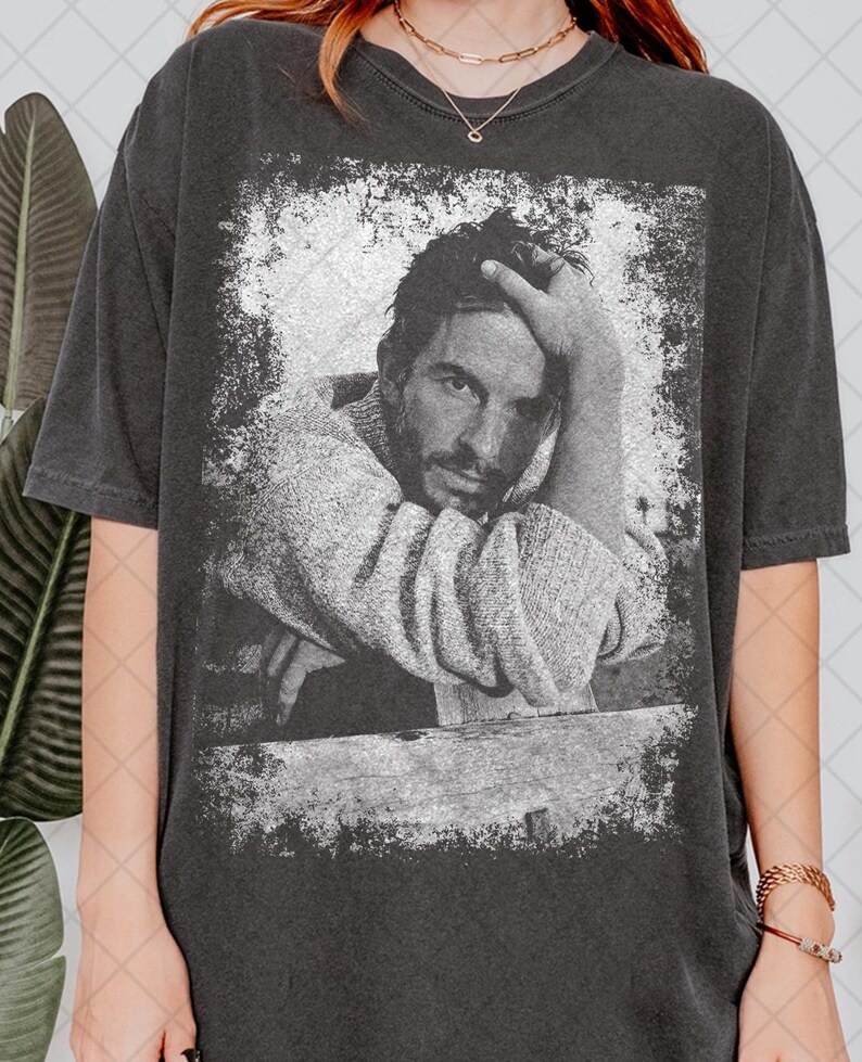 Puede incluir: Camiseta gris oscuro con una imagen en blanco y negro de una persona apoyando la cabeza en su mano. La imagen tiene un aspecto envejecido y vintage. La camiseta tiene mangas cortas y cuello redondo.