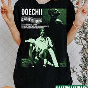 Könnte beinhalten: Schwarzes T-Shirt mit einem grünen und weißen Grafikdruck eines Rappers mit einem Alligator. Der Text auf dem Shirt lautet "DOECHII ALLIGATOR BITES NEVER HEAL".